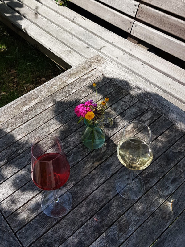 Ein Glas Weiß- und ein Glas Rotwein am Holztisch, der Tisch steht im Halbschatten unter einer Weinlaube